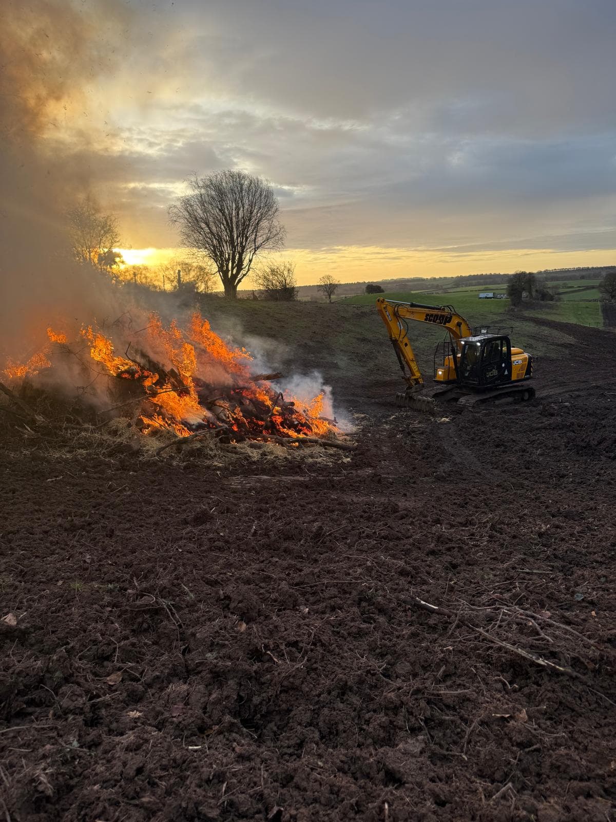 Excavator land clearance with bonfire at sunset